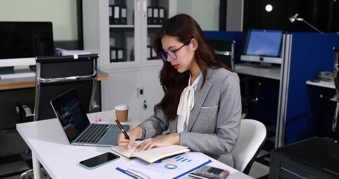 Young businesswoman in glasses working at office desk, writing notes beside laptop, phone and charts in a modern workspace.