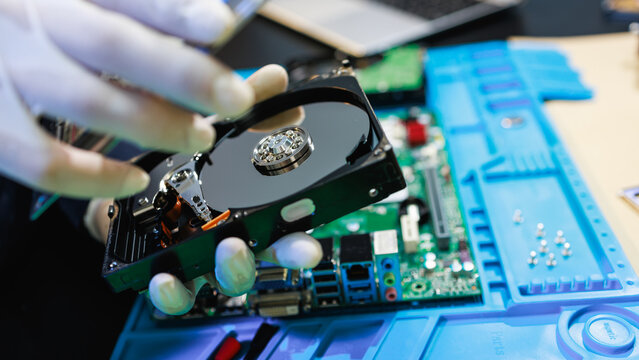 Technician in gloves repairing an open hard drive over a computer motherboard on a workbench, showing data recovery, hardware maintenance, and electronics service.