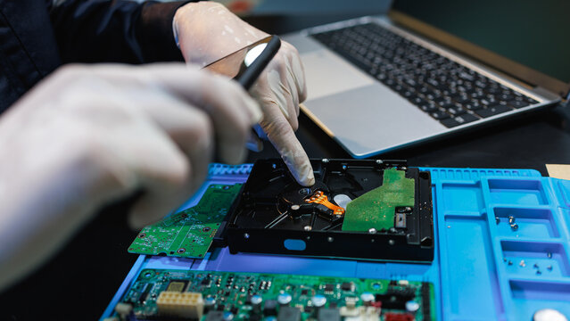 Technician in white gloves repairing an open hard drive on a workbench with circuit boards, laptop and tools, close up view for data recovery, electronics service and computer maintenance.