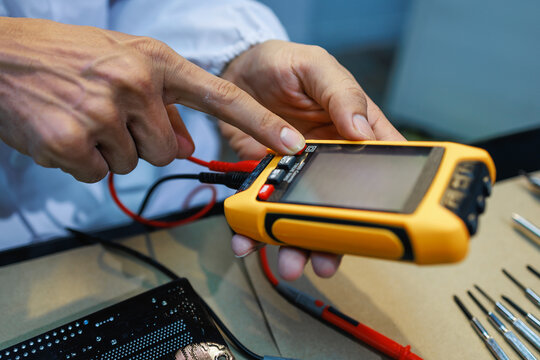 Close-up of a technician using a handheld digital tester with probe wires at a workbench, checking electronic equipment during repair or maintenance.
