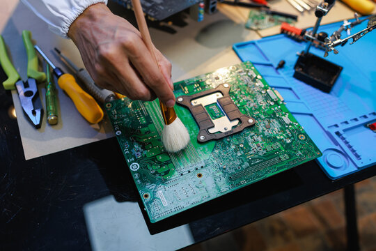 Close-up of a technician cleaning a computer motherboard with a soft brush on a repair desk with tools, showing electronics maintenance, hardware service and careful work.