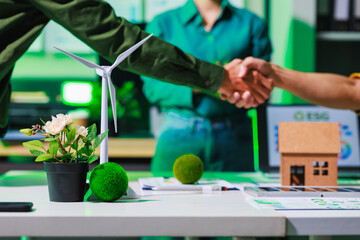 Business partners shake hands in a green office with a wind turbine model, small house and plant,...