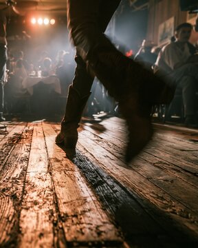 Midstep line dancing boots sharply framed against aged floorboards hazy crowd tables behind capturing motion blur and dramatic lighting that evoke lively country concert vibes.