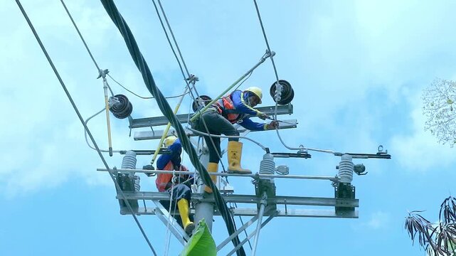 Two electricians work high on a power line tower. They wear safety gear and harnesses. Their tools and equipment hang nearby. Sky is blue with scattered clouds above. They focus on their task