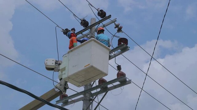 Utility workers maintain power lines on a sunny day. They work from a lifted bucket platform. Wearing helmets and safety gear, they focus on repairs. The sky shows blue with scattered clouds