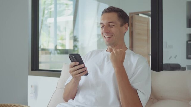 Financial success man smiling at smartphone after receiving positive notification and celebrating at home. Financial success concept reflects profit income online earning and digital finance lifestyle