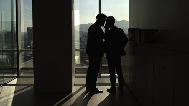 Silhouettes of two businessmen sharing a confidential secret by a large office window overlooking a city and mountains. Emphasizing discreet communication and privacy in business.
