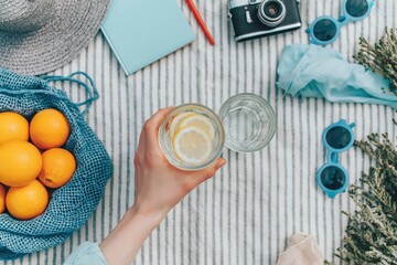 Fototapeta premium Picnic scene with hand holding refreshing lemonade glass, summer fruit, oranges, camera