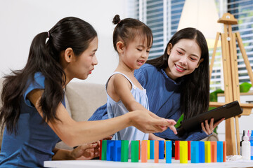 Obraz premium Two women and a young girl play with colorful blocks while looking at a tablet in a bright living room, showing family learning, bonding, and fun at home.