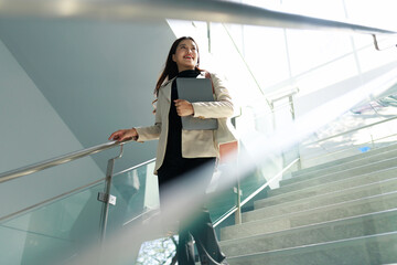 Smiling businesswoman walking down a bright modern office staircase, holding a laptop and looking confident in a clean professional setting. © Phushutter