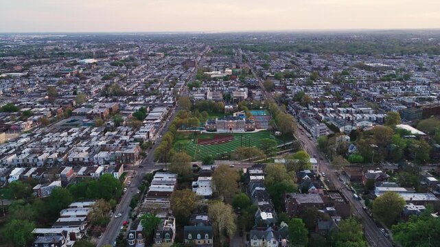 Wide aerial drone view of a vast Philadelphia rowhouse neighborhood at dusk with a school athletic field and building visible at the center of the cityscape.
