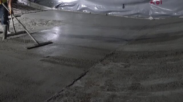 Construction workers in safety boots leveling fresh concrete over steel rebar grid during a night shift [cite: AA_17.jpg]. Detail shot of industrial slab pouring and site operations
