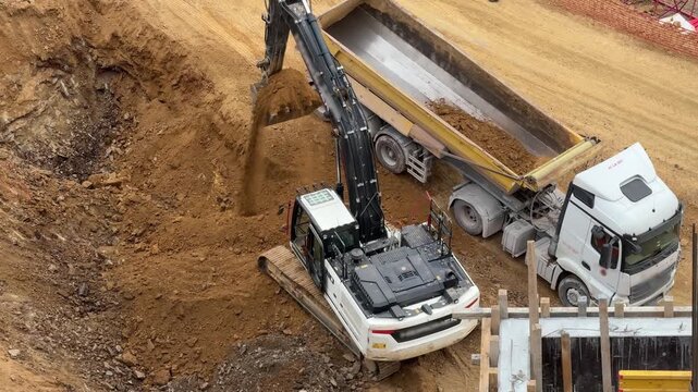 Crawler excavator loading soil and stones into the tipper of a white dump truck at an open construction site. Heavy machinery logistics field operation.