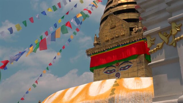 A static low-angle medium shot of the golden spire and wisdom eyes of Swayambhunath Stupa, framed by colorful prayer flags and a white shrine structure in Nepal.