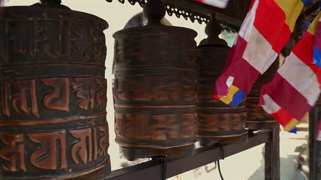 A close-up static shot of traditional bronze Tibetan prayer wheels spinning rapidly with colorful Buddhist flags fluttering in the background in Nepal.