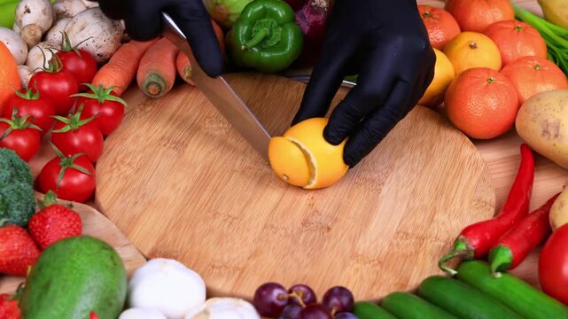 Chef in black gloves slicing a fresh lemon on a wooden cutting board surrounded by assorted vegetables and fruits.
