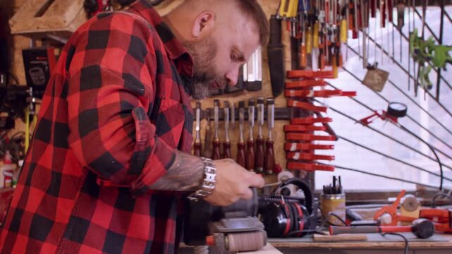 Lumber polishing. Carpentry tool. Male joiner in red red plaid shirt smoothing plank surface with professional electric equipment in workplace.