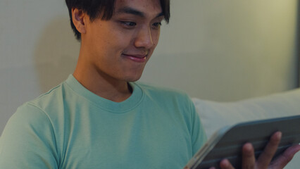 Close up young Asia male with casual cloth sit on sofa finger scroll tablet watch online movie in living room cozy house at night. E-book reading, Shopping online, Lifestyle leisure at home concept. © tirachard