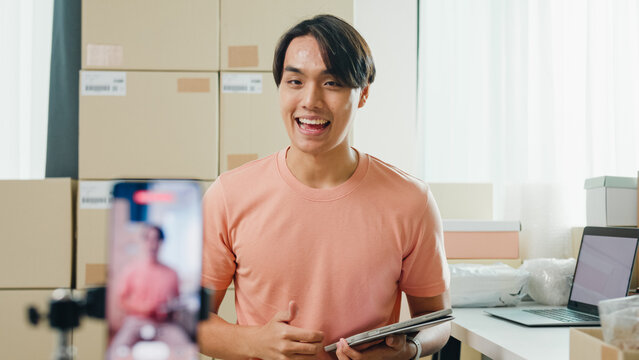 Youth attractive Asia male owner small business online shop sit front of phone look at camera live streaming see comment on digital tablet from customer sales promotion at retail online shop at home.