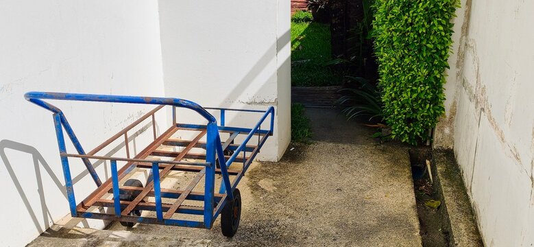 Old blue steel hand cart trolley parked outdoors near a white wall.