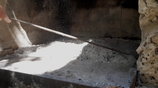 Worker uses long metal tool to gather and level hot ash in rustic stone oven area