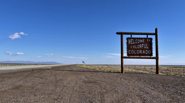 Atmospheric 4K timelapse of the iconic rustic Welcome to Colorful Colorado road sign along a desolate highway. Sweeping expansive plains and a vibrant blue sky create a cinematic travel aesthetic.