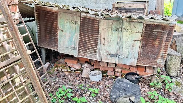 Weathered wooden storage shed elevated on stacked bricks in a neglected rural yard. Peeling paint, rusted shutters, and signs of decay highlight abandonment, aging structures, and rustic countryside l