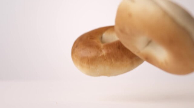 Chewy golden bagels dropping and bouncing across white seamless backdrop in high speed slow motion. breakfast catering posts, hotel buffet content, coffee shop pastry videos.
