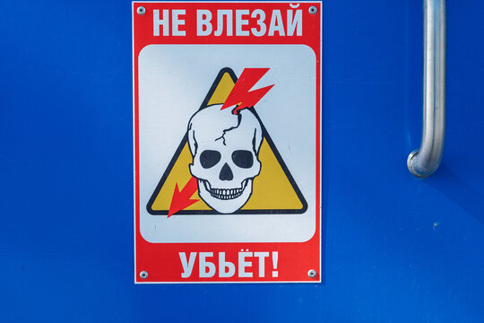 Warning sign displays a skull and lightning bolts, with Cyrillic text DON'T GET INVOLVED IT WILL KILL indicating danger, mounted on a blue surface for visibility