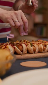 Vertical closeup of unrecognizable woman placing small American flags into hot dogs on wooden board. Footage is perfect for Fourth of July party planning and holiday food marketing