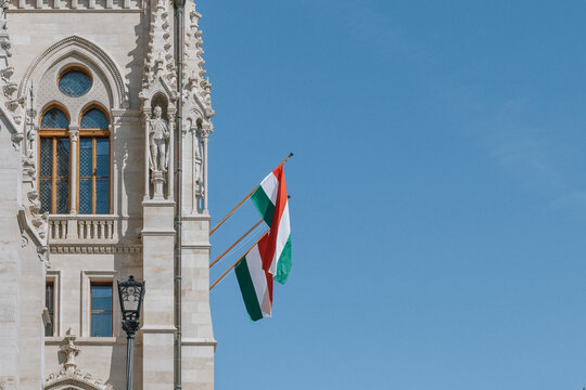 Hungarian national flags mounted on an ornate historic stone facade with arched windows, decorative sculpture and traditional street lamp under a clear blue sky