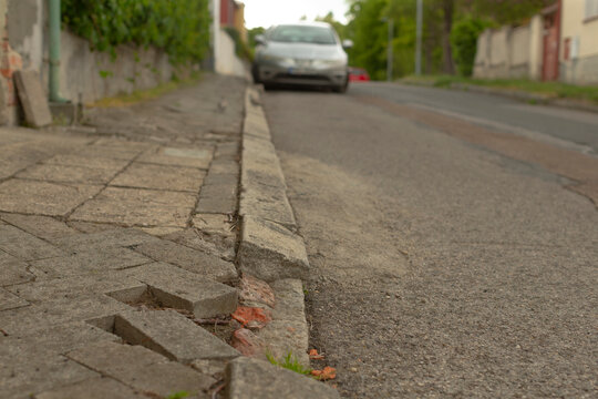 Close low-angle view of a deteriorated concrete curb and uneven paved sidewalk next to an asphalt residential road. Foreground shows broken paving blocks