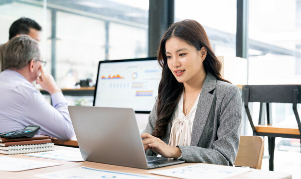 Asian businesswoman working on laptop with corporate team in background.