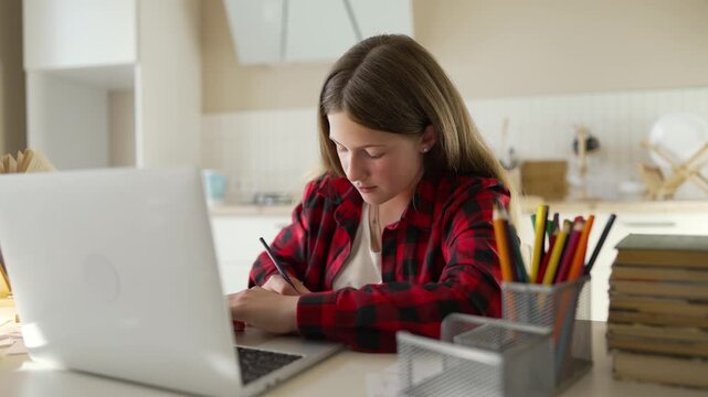 Teenager sits at desk at home. Focused student is studying with open laptop. Girl writes down important homework notes. Diligent student looks at bright laptop screen. Studying requires focus.