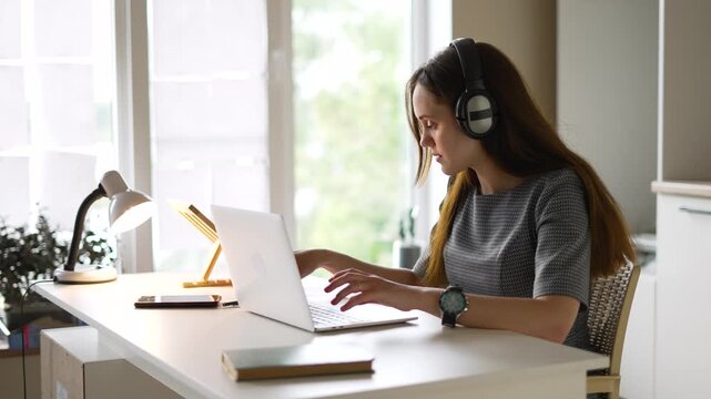 Young woman typing on modern laptop keyboard. Focused student working inside bright home office. Woman wearing large headphones studying. She sits typing at desk. Daylight shines near laptop.