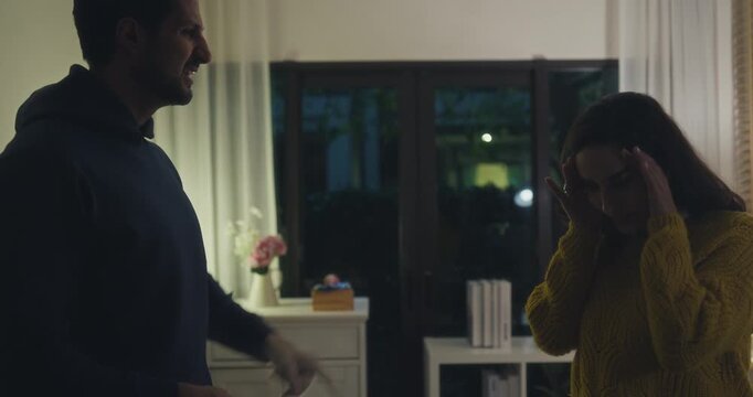 Angry Hispanic young couple arguing and pointing fingers in dark home living room at night. Domestic conflict dispute, toxic relationship problem, divorce crisis, communication breakdown concept.