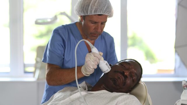 Middle-aged African American undergoing hardware facial treatment using cold hammer helping to shrink pores, repair damaged skin and relieve inflammation in modern aesthetic medicine clinic