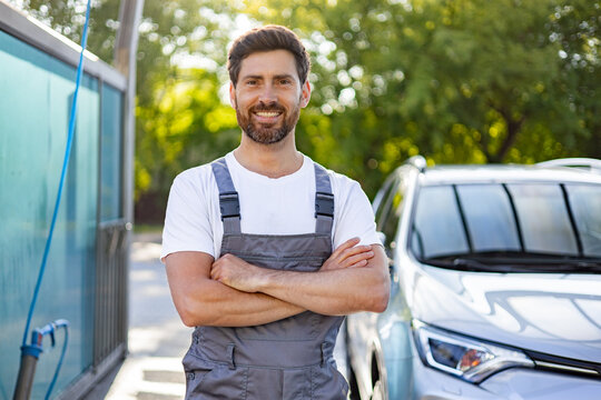 A smiling Caucasian man in overalls stands with his arms crossed in front of a car at a car wash