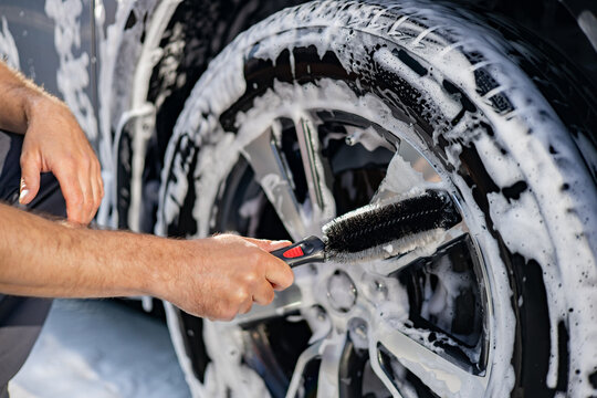 A person's Caucasian hands meticulously scrub a soapy car wheel with a brush, highlighting the detailed cleaning process