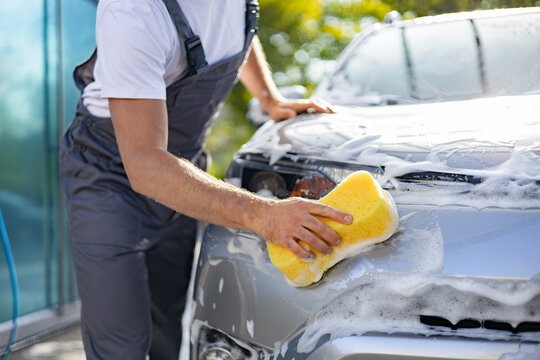 A man in overalls washes a car with a yellow sponge and plenty of foam, outdoors on a sunny day