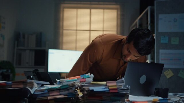 Stressed young Latin businessman walking to place tax document stack at desk in dark night office. Overworked overtime, heavy workload stress, tired employee, corporate business burnout concept.