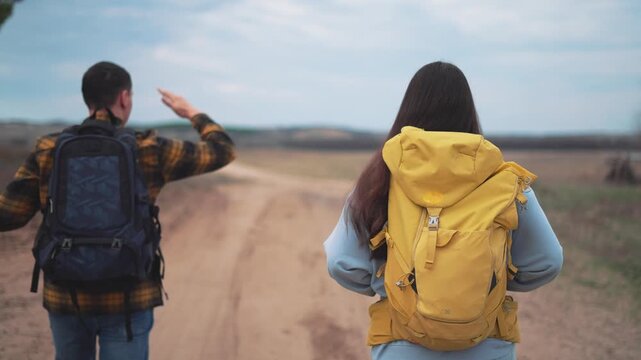 Couple backpackers walking on desert road pointing forward during adventure travel journey. Man and woman with backpacks hiking empty trail. Travelers explore wilderness path together on trip.