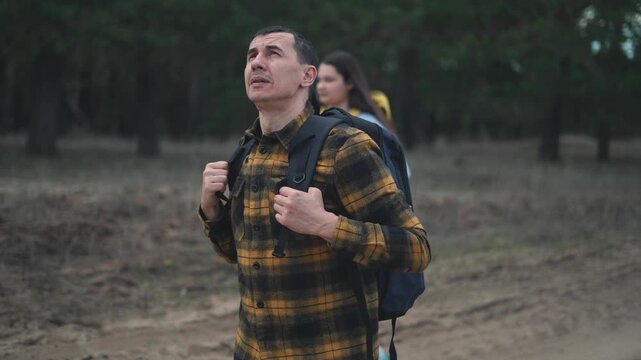 Adventurous man hiking with black backpack explores forest trail. Male hiker in yellow plaid shirt gazes upward during nature trek. Outdoor enthusiast enjoys wilderness journey on dirt path.