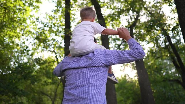 Father carries son on shoulders walking through sunny park. Happy dad lifts child high outdoors. Parent and boy bonding in nature together. Man carrying kid among green trees in summer forest.
