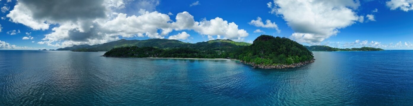 Panoramic drone view of Mah&eacute;'s lush coastline from Anse Louis to Anse La Mouche, Seychelles, featuring vibrant tropical mountains, blue Indian Ocean waters, and dramatic skies