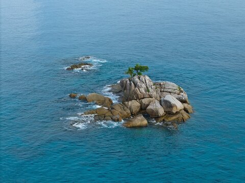 Aerial view of a lonely rocky islet with green trees surrounded by deep blue and turquoise ocean waters, Anse solei, Mahe, Seychelles 