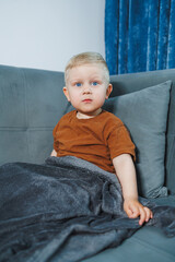 Fototapeta premium Blond caucasian toddler calm direct gaze into camera, neutral expression, wrapped in gray blanket, orange tee, soft living room environment, intimate childhood portrait