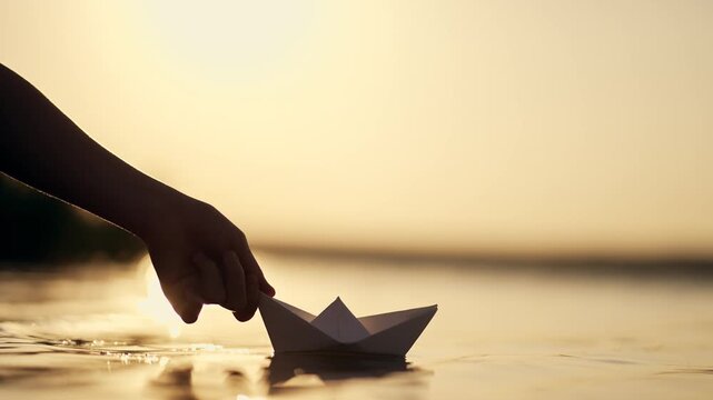 Silhouetted hand gently placing white paper origami boat on calm water surface during golden sunset. Beautiful warm evening light creates soft reflections and peaceful atmosphere by the shore.