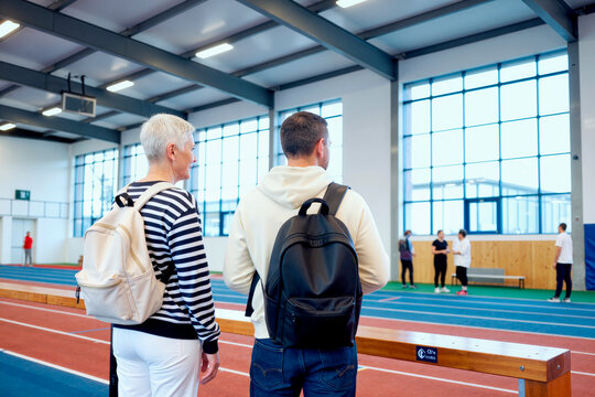 Senior Caucasian woman with young adult man standing beside indoor running track, observing training session and discussing fitness program. Useful for active aging or sports education marketing