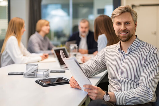 Smiling handsome businessman manager sitting on the table in office and reading documents and looking at camera, colleagues on the background